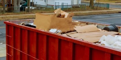 A red dumpster filled with cardboard boxes and various discarded items in a neighborhood setting.