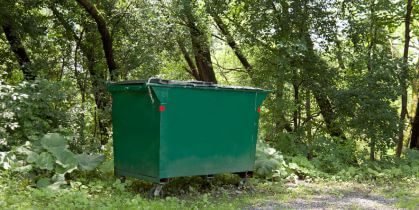 Green dumpster in a wooded area surrounded by trees and foliage.