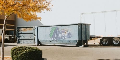 Grey Dumpster A large blue dumpster with a truck illustration stands proudly next to a white trailer, framed by an autumn tree and lush green shrubbery.